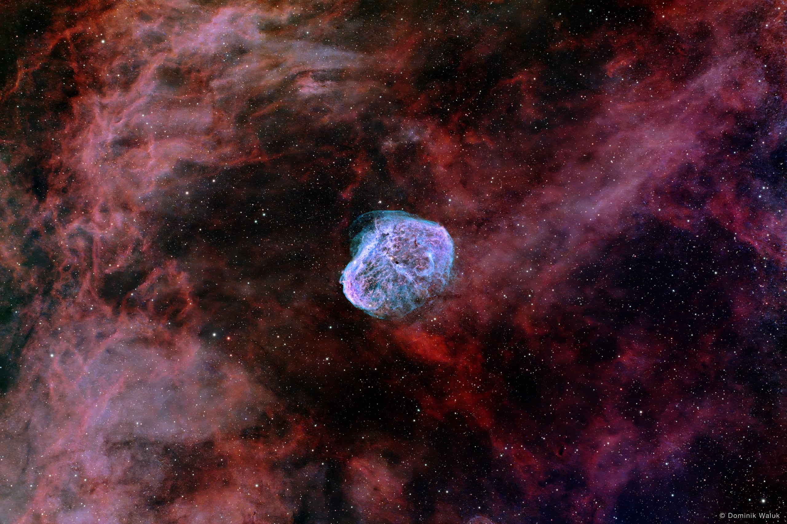 Der Mondsichelnebel: eine blau schimmernde, schalenförmige Wolke mit feinen Filamenten inmitten dichter roter Nebel.